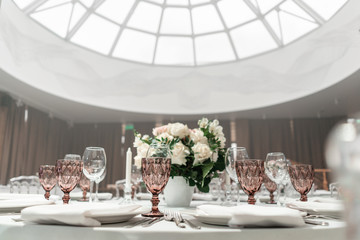 Wine glasses on round Banquet table served. Interior of restaurant for wedding dinner, ready for guests. Decorated with floral arrangement. Dishes, wine glasses and napkins. Catering concept.