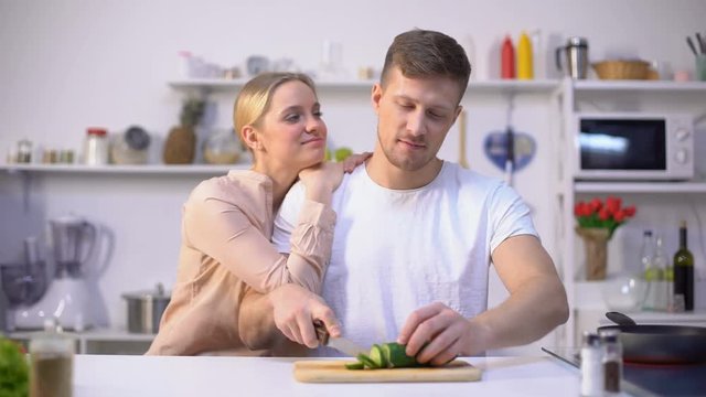 Husband slicing vegetable, loving wife embracing him, romantic moment in kitchen