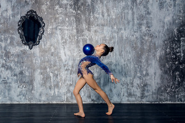 Girl gymnast in a blue suit makes exercise with a ball against a gray wall.