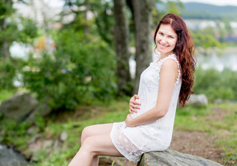 Fototapeta premium Portrait of cute young caucasian woman in nice white dress in the summer park. Pregnant model sits on a stone and holds her stomach.
