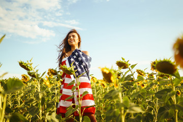 Obraz na płótnie Canvas Beautiful girl with the American flag in a sunflower field. 4th of July. Fourth of July. Freedom. Sunset light The girl smiles. Beautiful sunset. Independence Day. Patriotic holiday.