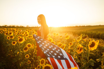 Obraz na płótnie Canvas Beautiful girl with the American flag in a sunflower field. 4th of July. Fourth of July. Freedom. Sunset light The girl smiles. Beautiful sunset. Independence Day. Patriotic holiday.