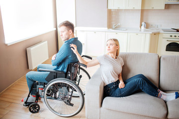 Fototapeta na wymiar Young man with special needs sit on wheelchair back to back with woman. She touch his shoulder with hand and look at him. Upset and unhappy couple.