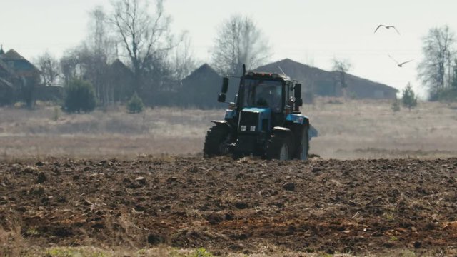 Static shot of a blue tractor with big black wheels and a powerful grouser, heavy plowing the dark fertile soil. The farm machine is followed by white birds. The warmth from the ground and the motor