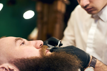 Cutting mustache with sharp scissors. Barber in black hygienic gloves concentrate in scrupulous work process. Mustache styling and cutting. Advertising for barber shop. Side view. Blurred background.