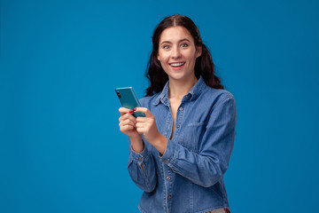 Fototapeta premium Young brunette woman is posing with a smartphone standing on blue background.