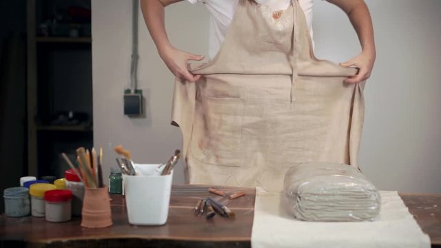 Woman is putting on an apron in her handicraft workshop, standing near table,