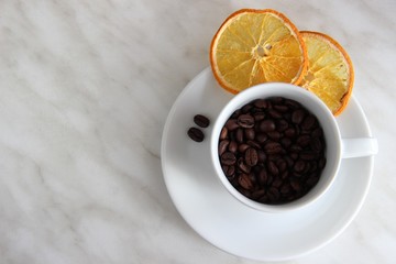 One Cup and saucer filled with roasted coffee beans, with two slices of dried orange and a pair of coffee beans on the background of gray table top