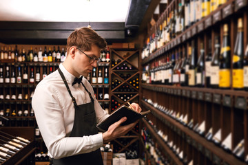 Professional male Sommelier dressed in bowtie and apron writes at notepad, putting down information on wine bottles in wine house