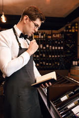 Thoughtful male sommelier looking at a very wide range of wines from the main regions of the world, writing in note pad some things working at wine restaurant.