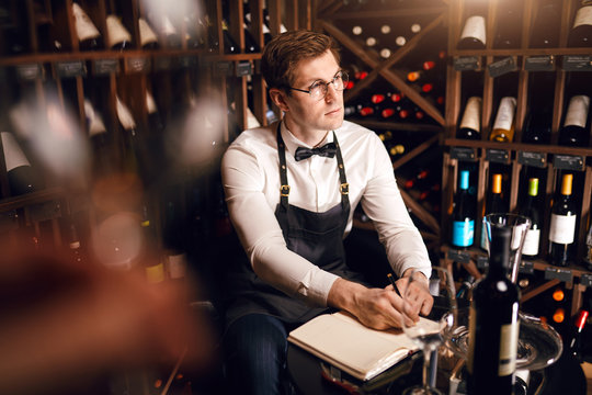 Blurred shot of pensive man sommelier writing in note pad some things sitting between a very wide range of wines placed on shelves at wine restaurant.