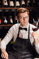 Bartender or male cavist standing near the shelves of wine bottles holds a glass of wine, looks at tint and smells flavor of wine in glass.