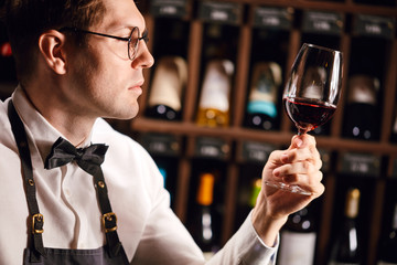 Bartender or male cavist standing near the shelves of wine bottles holds a glass of wine, looks at tint and smells flavor of wine in glass.