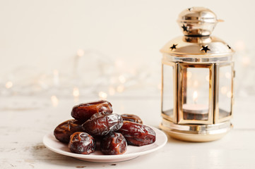 Dried dates on white plate and lantern with burning candle on white wooden table, bokeh light on background. Ramadan concept