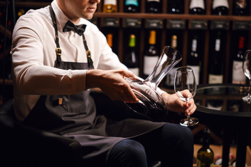 Cropped view of professional sommelier pouring red wine from bottle into decanter at table in wine restaurant.