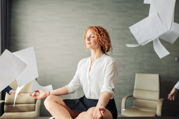 Calm young red-haired woman in formal wear having yoga meditation on table in office being surrounded with flying papers. Taking a break for relaxation. Work and recreation concept.