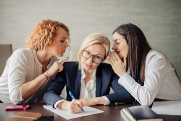 Blonde business woman working, writing a document and two female coworkers gossiping in her ears...