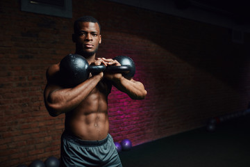 Young ripped African male athlete with perfect well-shaped body posing with dumbbells on brick wall background.