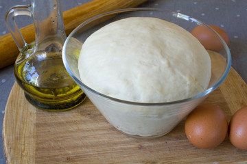 Fresh raw dough in a glass bowl on a wooden board with eggs and oil.