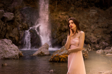 Attractive girl posing on the rock among in fairy fast forest in autumn and beside beautiful waterfall with blue water. Fairy tale.