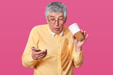 Astonished old man poured coffe on his yellow shirt while reading shocking information in his smart phone, wears glasses, has surprised facial expression, poses over pink studio wall, empty space.