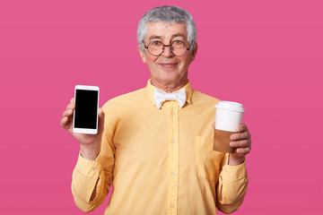 Close up portrait of mature grey haired man in yellow shirt with bow tie, holds mobile phone with blank screen and takeaway coffee, looks directly at camera, isolated over pink studio background.