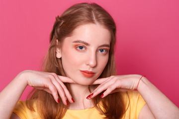 Romantic tender young lady holds fingers with red manicure near cute fresh face with make up posing over dark pink background in studio, having red thread on her wrist, looks sweet and pure.