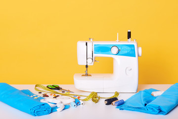 Studio shot of sewing equipments, sewing machine, tap measure, scissors, pieces of cloth, needles, thread isolated over yellow studio background. Image of different tools in sewing workshop,