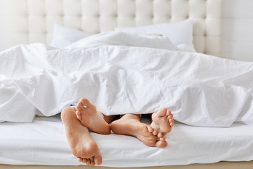 Horizontal shot of feet of couple under white bedclothes in bed at bedroom, enjoying good rest at home and calm atmosphere. Female and male relaxing together at weekend. Pomantic couple in love.