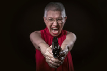 Screaming frustrated senior old woman in a red dress aiming a gun at the camera