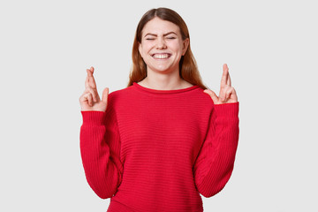 Indoor shot of pleasant looking dressed in casual red sweater, crosses fingers and has great hope for better, waits results of examination, posing in studio with cluosed eyes and toothy smile.