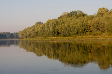 Wide river flowing across green forest. Fall. Reflections of sun in the water