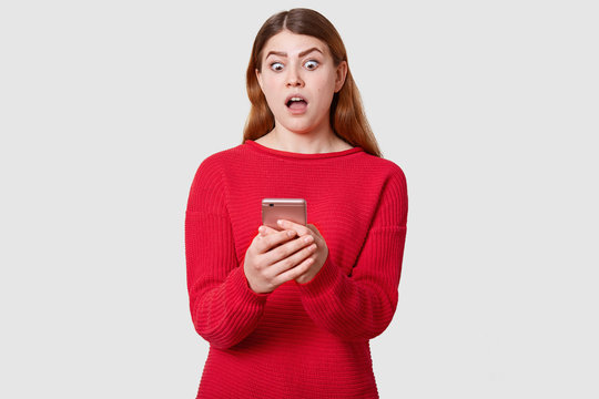 Front view of surprised model holds her smartphone with both hand, looking at display with astonished facial expression, has shoking news fron friend, reading message, isolated over white background.