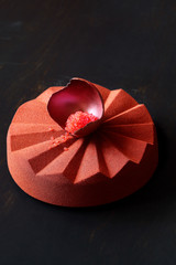 Contemporary Chocolate Mousse Cake made in geometric silicone mold, covered with brick color velvet spray and decorated with chocolate shell filled with blood orange caviar, on black background.