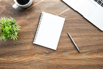 Blank notebook page is on top of wood office desk table with laptop computer, cup of coffee and supplies. Top view with copy space, flat lay.