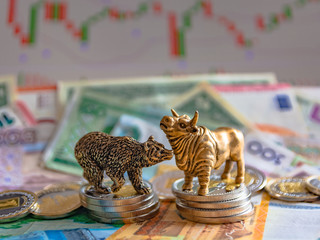 Bronze figures of a bull and a bear near metal coins against the background of paper money and charts. Blur background and perspective. Concept and symbol of stock exchange and stock trading.