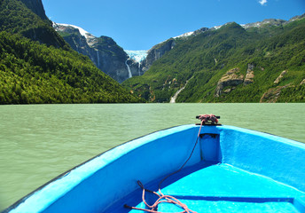 Front of a small blue boat in the light green icy waters of Ventisquero lagoon, on a sunny summer morning, going for a tour, with Ventisquero Colgante and its waterfalls on the background