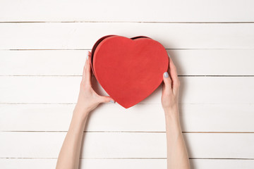 Female hands hold a red heart shaped box against the background of a white wooden table