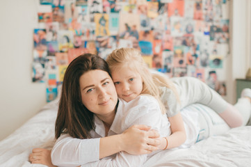 Lifestyle soft focus portrait of happy mom hugs her adorable young daughter on white bed. Morning family portrait of smiling mother and her funny daughter hugs on 90s style wall on background.