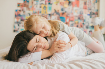 Obraz na płótnie Canvas Lifestyle soft focus portrait of happy mom hugs her adorable young daughter on white bed. Morning family portrait of smiling mother and her funny daughter hugs on 90s style wall on background.