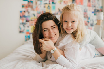 Lifestyle soft focus portrait of happy mom hugs her adorable young daughter on white bed. Morning family portrait of smiling mother and her funny daughter hugs on 90s style wall on background.