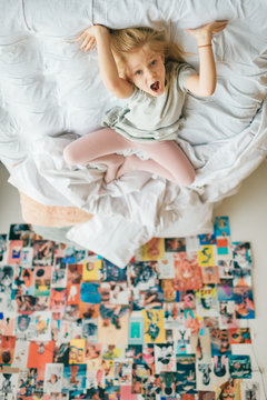 Funny little girl clowning, fooling and make faces on white bed  in child room. Cheerful young female kid lying on bed with emotional feelings on her face and 90s style decorative wall on background