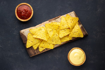 Mexican corn chips nachos with sauces on rustic cutting board. Dark background. Top view.