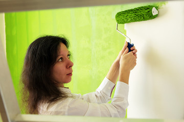 Pretty young woman in a white shirt is carefully painting green interior wall with roller in a new home