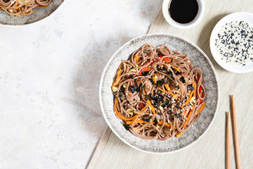 Buckwheat noodles with vegetables and meat decorated with sesame and seaweeds in bowls on grey background.