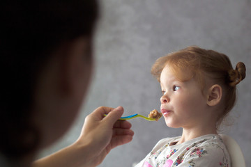 mother feeds her daughter with a small spoon, mother gives food to her red-haired little girl