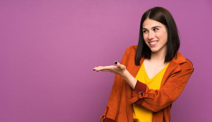 Young woman with a coat over isolated purple background extending hands to the side for inviting to come
