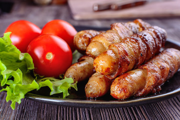 Appetizing sausages wrapped in bacon and grilled next to tomatoes and lettuce are on a wooden table