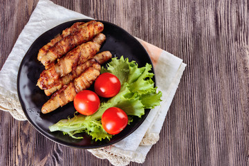 Appetizing sausages wrapped in bacon and grilled next to tomatoes and lettuce are on a wooden table, top view