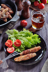 Appetizing sausages wrapped in bacon and grilled next to tomatoes, lettuce and and glass of beer are on a wooden table, vertical